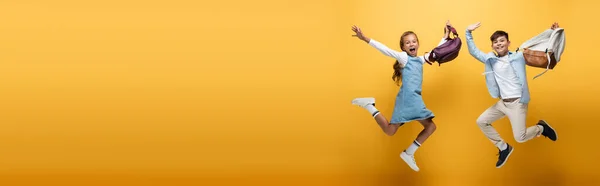 Excited interracial schoolkids with backpacks jumping on yellow background, banner — Stock Photo