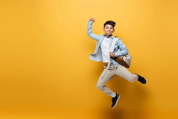 Asian schoolkid holding backpack and showing yes gesture while jumping on yellow background — Stock Photo