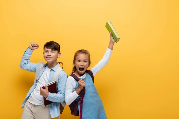 Aufgeregte interrassische Schulkinder halten Bücher in der Hand und zeigen Ja-Geste auf gelbem Hintergrund — Stockfoto