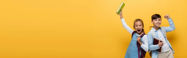 Excited multiethnic schoolkids holding books and looking at camera on yellow, banner — Stock Photo