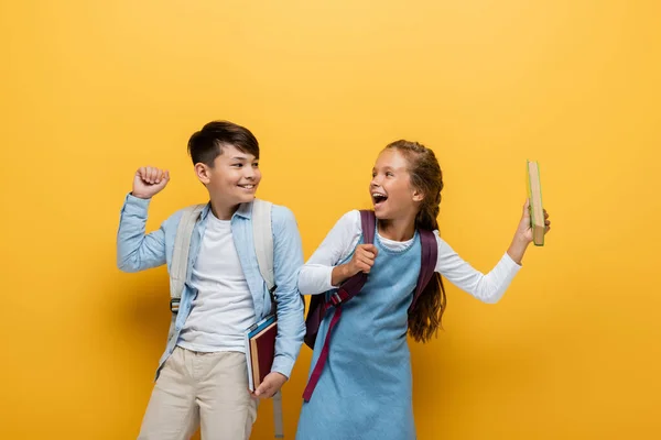 Entusiasmados alunos inter-raciais com mochilas e livros olhando uns para os outros em fundo amarelo — Fotografia de Stock