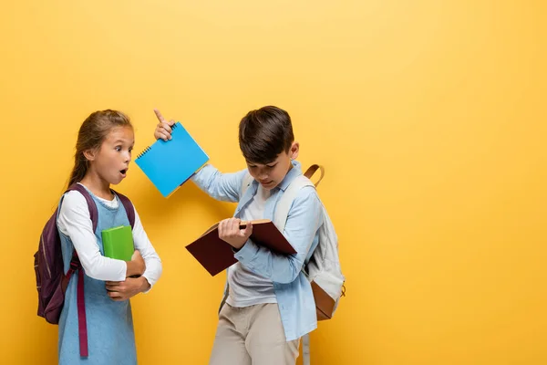 Shocked schoolgirl with book looking at asian friend with notebook and backpack isolated on yellow — Stock Photo