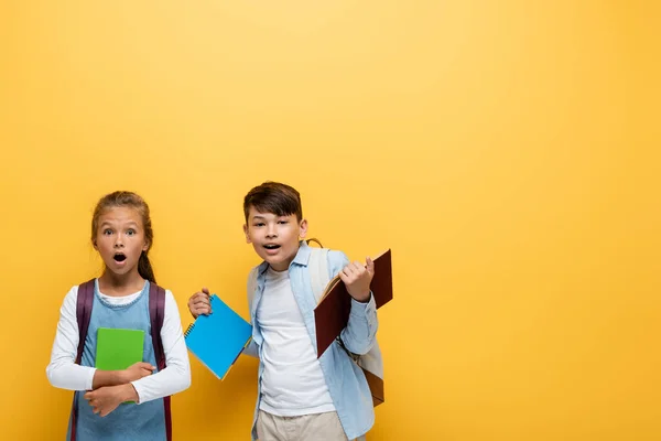 Sorprendidos escolares multiétnicos sosteniendo libros y mirando a la cámara sobre un fondo amarillo - foto de stock