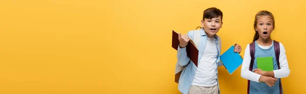 Compañeros de clase interracial impactados sosteniendo libros y mirando a la cámara sobre fondo amarillo, pancarta - foto de stock