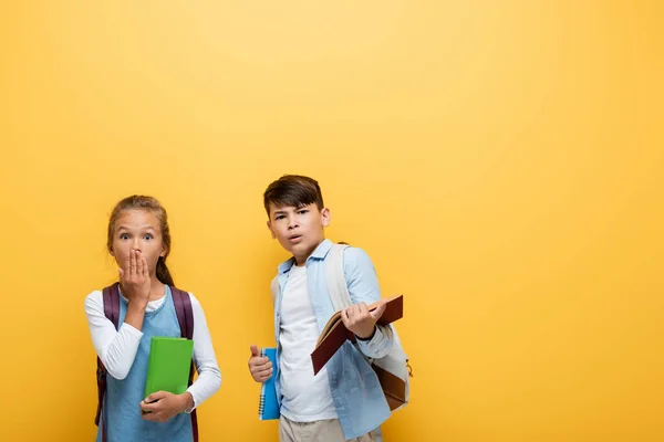 Shocked interracial pupils with books looking at camera isolated on yellow — Stock Photo