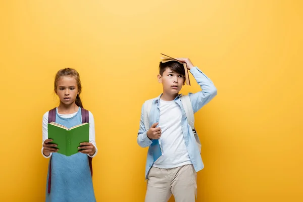 Asian schoolkid holding book near head and classmate isolated on yellow — Stock Photo
