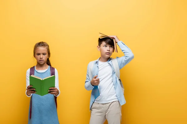 Asiático estudante segurando livro perto da cabeça e colega de classe isolado no amarelo — Fotografia de Stock