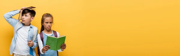 Asiático escolar sosteniendo libro cerca de la cabeza mientras amigo leyendo aislado en amarillo, bandera - foto de stock