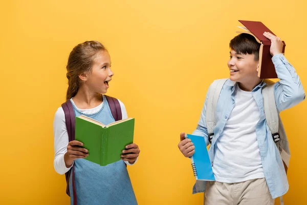 Positive interracial kids holding books and looking at each other isolated on yellow — Stock Photo