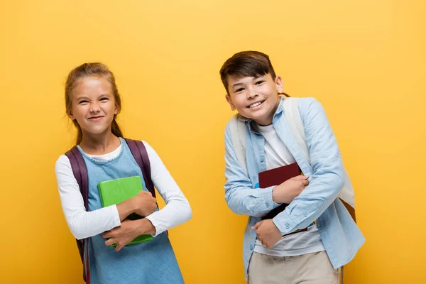 Colegiales multiétnicos sosteniendo libros y mirando a la cámara aislada en amarillo — Stock Photo