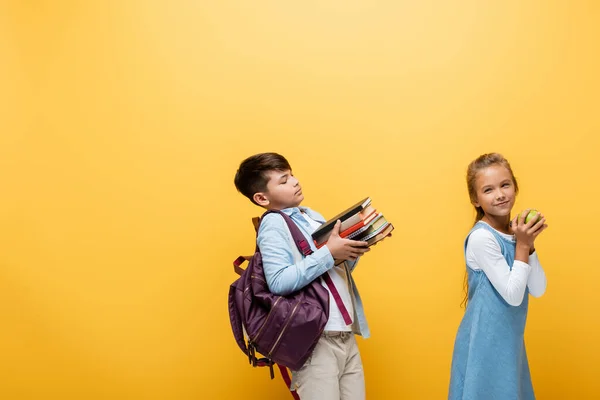 Asiatico scolaro con zaino tenendo libri vicino amico con mela isolato su giallo — Foto stock