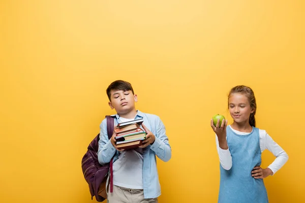 Asiático escolar celebración libros cerca de compañero de clase con manzana aislado en amarillo - foto de stock