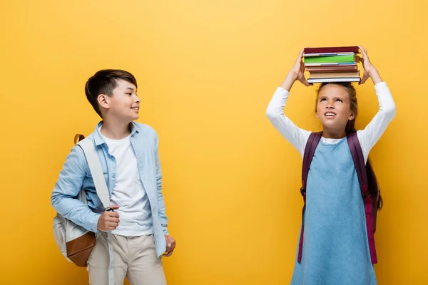 Smiling asian schoolboy holding backpack near friend with books isolated on yellow — Stock Photo