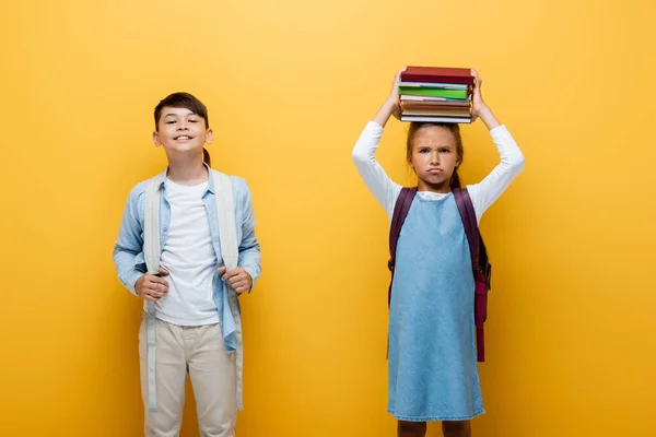 Smiling asian schoolboy holding backpack near upset friend with books isolated on yellow — Stock Photo