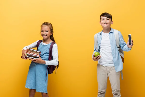 Alegre colegiala sosteniendo libros cerca asiático amigo con manzana y smartphone aislado en amarillo - foto de stock