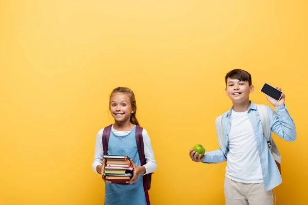 Escolares interraciales sonrientes sosteniendo libros y teléfonos inteligentes aislados en amarillo - foto de stock