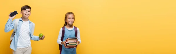 Allegro scolari multietnici in possesso di libri e cellulare isolato su giallo, banner — Foto stock