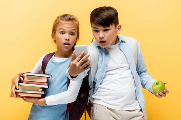 Shocked interracial schoolchildren with apple and books looking at smartphone isolated on yellow — Stock Photo