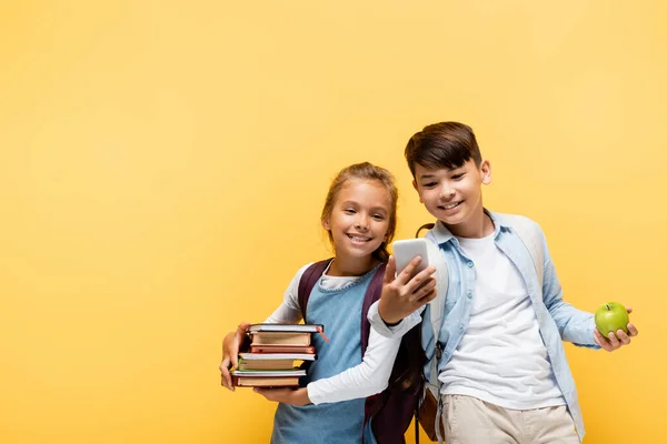 Souriant élèves multiethniques avec des livres et de la pomme à l'aide d'un téléphone portable isolé sur jaune — Photo de stock