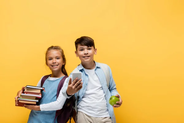Весёлые межрасовые школьники держат книги и смартфон изолированными на жёлтом — стоковое фото