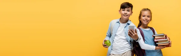 Preteen interracial schoolkids holding books and mobile phone isolated on yellow, banner — Stock Photo
