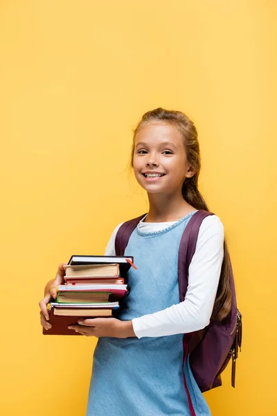 Estudante sorridente com mochila segurando livros isolados em amarelo — Fotografia de Stock