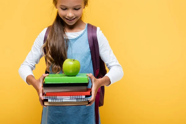 Усміхнена школярка дивиться на книги та яблуко ізольовано на жовтому — стокове фото