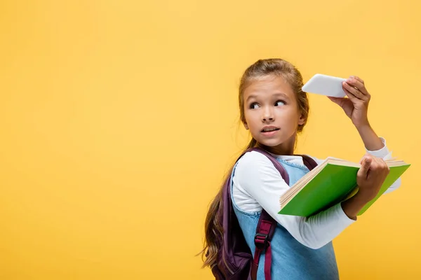 Colegial sosteniendo teléfono inteligente y libro aislado en amarillo - foto de stock