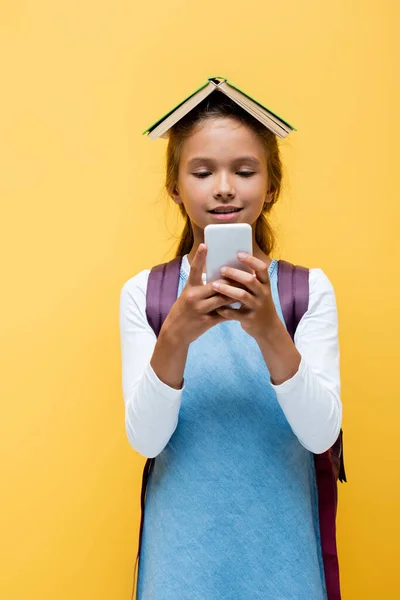 Escolar sonriente con libro en la cabeza usando teléfono móvil aislado en amarillo - foto de stock