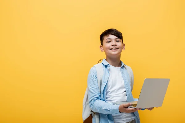 Souriant asiatique écolier tenant ordinateur portable isolé sur jaune — Photo de stock