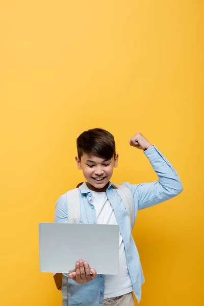 Aufgeregt asiatische Schulkind zeigt ja Geste und hält Laptop isoliert auf gelb — Stockfoto