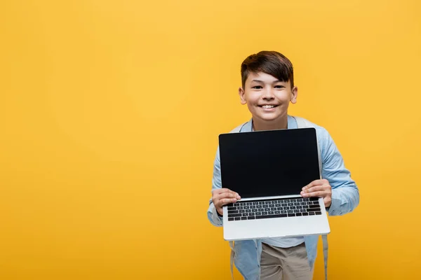 Sonriente asiático escolar sosteniendo portátil con pantalla en blanco aislado en amarillo - foto de stock