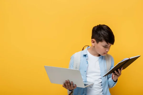 Asian schoolkid holding laptop and looking at notebook isolated on yellow — Stock Photo