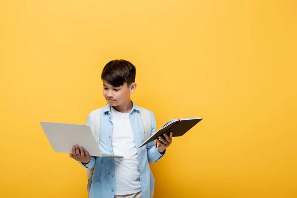 Asian schoolkid holding laptop and notebook isolated on yellow — Stock Photo