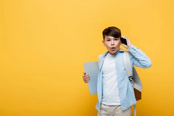 Shocked asian schoolboy talking on smartphone and holding laptop isolated on yellow — Stock Photo