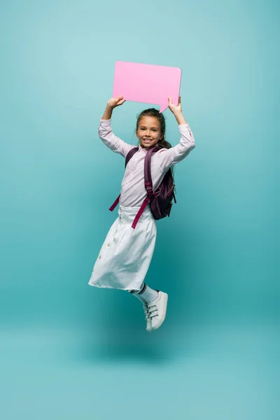 Positive pupil with backpack holding speech bubble on blue background — Stock Photo