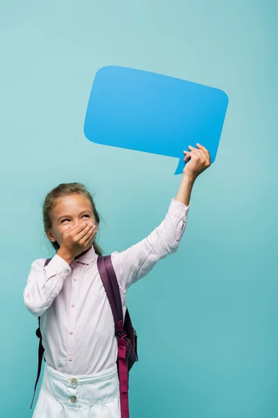 Laughing schoolgirl with backpack holding speech bubble isolated on blue — Stock Photo