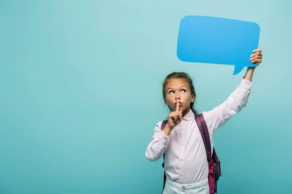 Schoolgirl with backpack holding speech bubble and showing secret gesture isolated on blue — Stock Photo