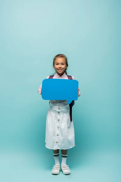 Positive schoolchild with backpack holding speech bubble on blue background — Stock Photo