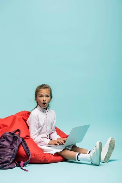 Escolar impactada usando el ordenador portátil en la silla del bolso de frijoles sobre fondo azul - foto de stock