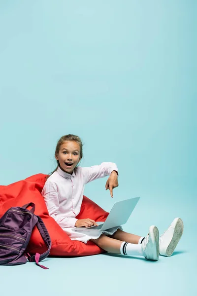 Écolier positif pointant avec le doigt à l'ordinateur portable près du sac à dos sur fond bleu — Photo de stock