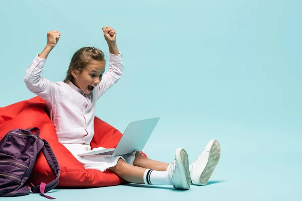 Excited pupil showing yes gesture while looking at laptop near backpack on blue background — Stock Photo