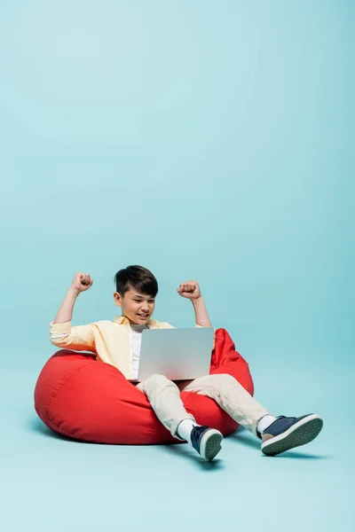 Excited asian schoolboy looking at laptop on beanbag chair on blue background — Stock Photo