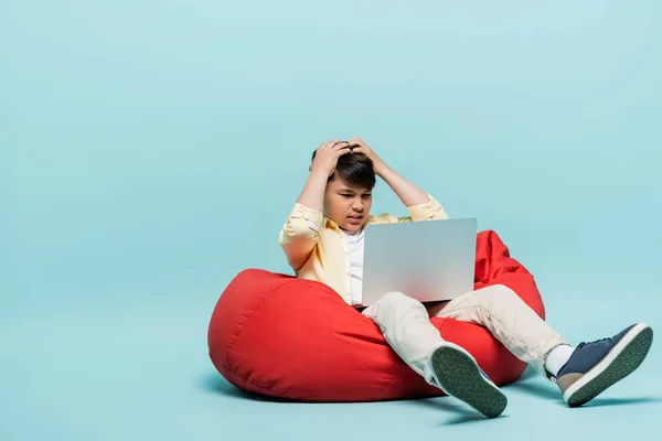 Worried asian schoolkid looking at laptop on beanbag chair on blue background — Stock Photo