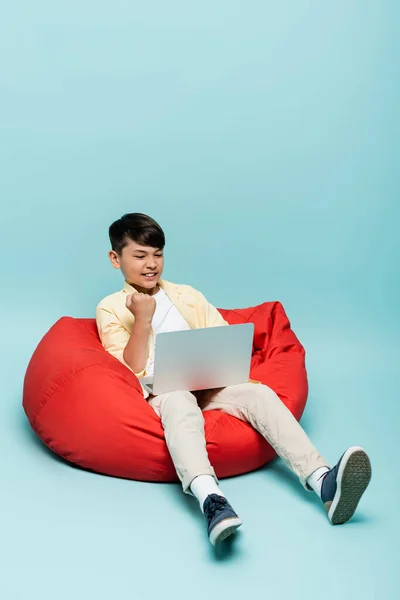 Excité asiatique écolier montrant oui geste tout en utilisant ordinateur portable sur chaise de haricot sur fond bleu — Photo de stock