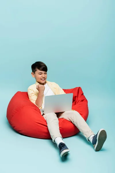 Emocionado asiático estudante mostrando sim gesto ao usar laptop na cadeira beanbag no fundo azul — Fotografia de Stock