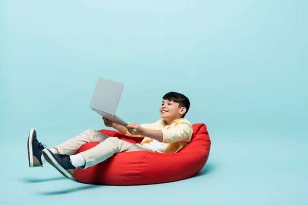Positive asian schoolkid holding laptop while sitting on beanbag chair on blue background — Stock Photo