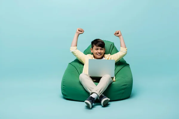 Asian schoolkid showing yes gesture while looking at laptop on beanbag chair on blue background — Stock Photo