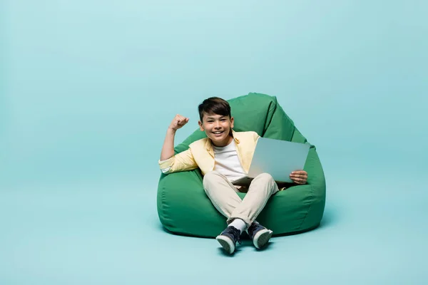 Smiling asian schoolkid showing yes gesture and holding laptop on beanbag chair on blue background — Stock Photo