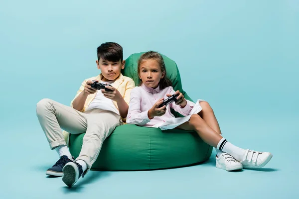 KYIV, UCRANIA - 2 de julio de 2021: Niños interracial enfocados jugando videojuegos en una silla de frijoles sobre fondo azul - foto de stock